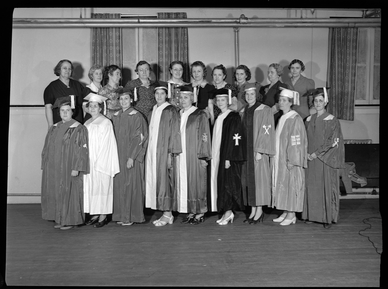 Nineteen women stand together in two rows. The women in the front row are all wearing graduation caps and gowns, while the women in the row behind them are not. They are all standing together in a room.