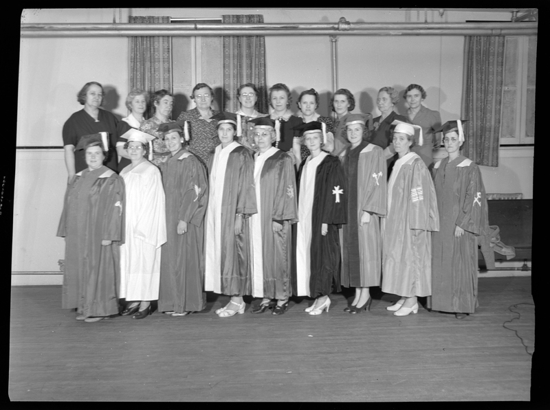 Nineteen women stand together in two rows. The women in the front row are all wearing graduation caps and gowns, while the women in the row behind them are not. They are all standing together in a room.