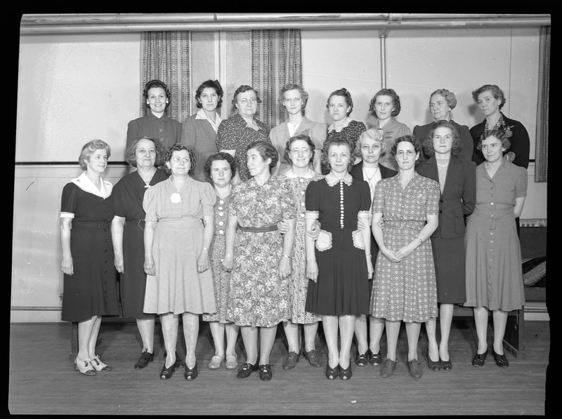 Nineteen women standing together in a room in two rows. Most of the women are looking at the camera.