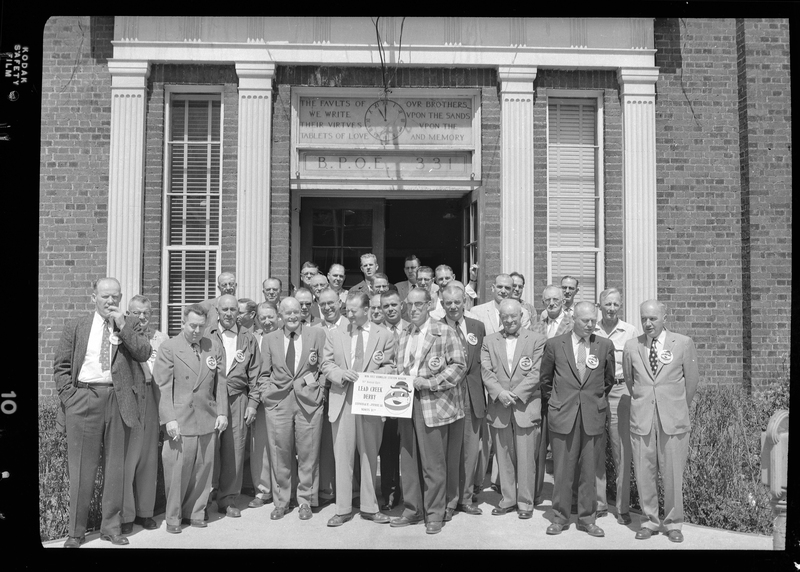 Group of men from the Gyro Club stand posed together in front of the Benevolent and Protective Order of Elks building. Two of the men are holding a sign up for the Lead Creek Derby and everyone is wearing matching buttons. The building's doors are open and you can see the clock that hangs above it.