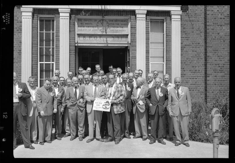 Group of men from the Gyro Club stand posed together in front of the Benevolent and Protective Order of Elks building, with one of the men cut off. Two of the men are holding a sign up for the Lead Creek Derby and everyone is wearing matching buttons. The building's doors are open and you can see the clock that hangs above it.