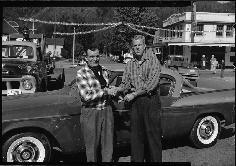 Two men from the Gyro Club stand in front of a car while shaking hands and looking at the camera. A street scene is visible in the background with cars, buildings, trees, and a few people. 