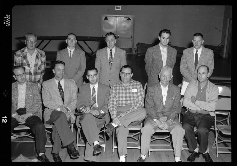 A group of men together in a room, half of which are sitting in a row of chairs while the other half stand behind them, all posing for a photo together. 