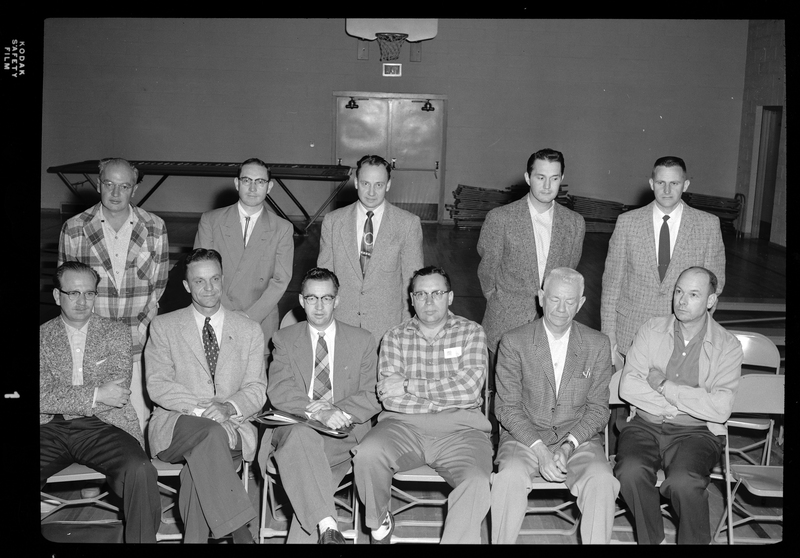 A group of men together in a room, half of which are sitting in a row of chairs while the other half stand behind them, all posing for a photo together. There is a basketball hoop visible on the back wall.