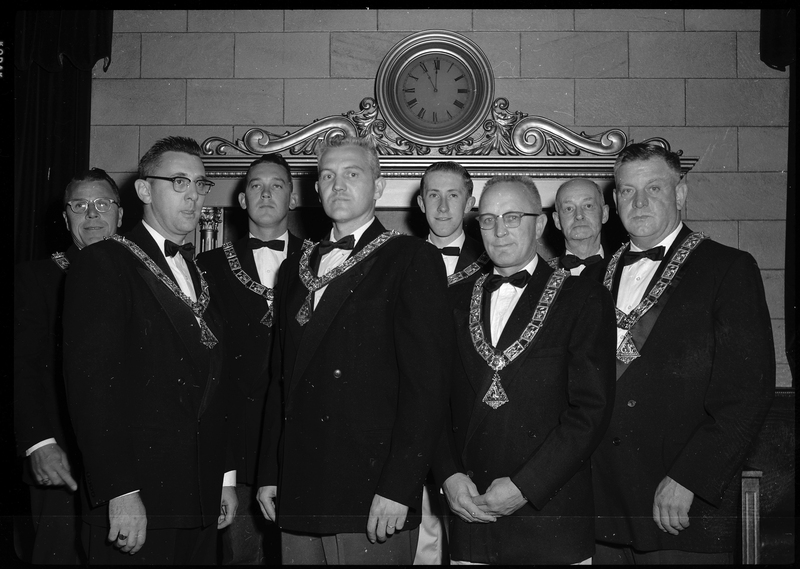 Photo of the eight men that are officers for the Benevolent and Protective Order of Elks. They are all wearing nice suit jackets and matching pendants while making serious faces. Most of the men are looking at the camera with a couple looking other places.