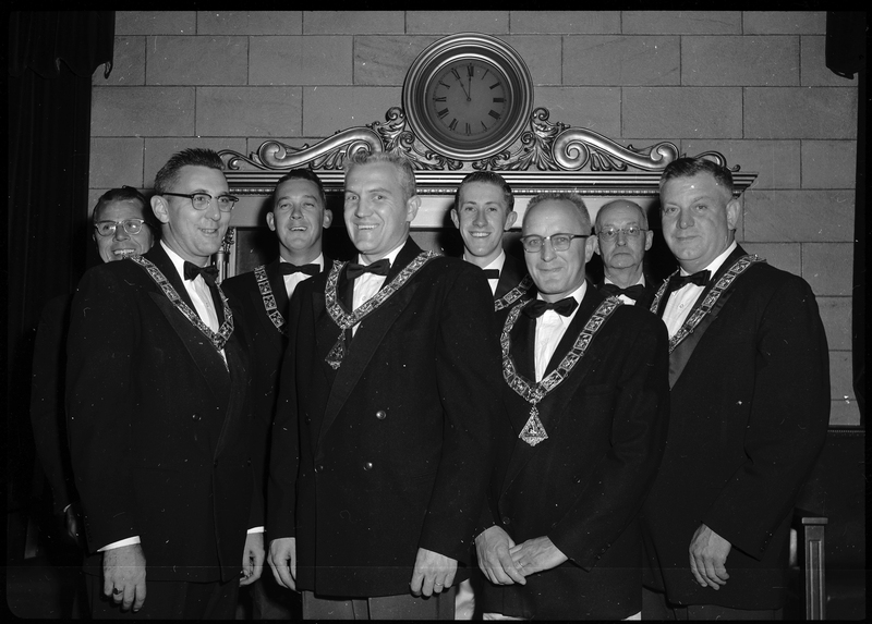 Photo of the eight men that are officers for the Benevolent and Protective Order of Elks. They are all wearing nice suit jackets and matching pendants and most are smiling. Most of the men are looking at the camera with a couple looking other places.