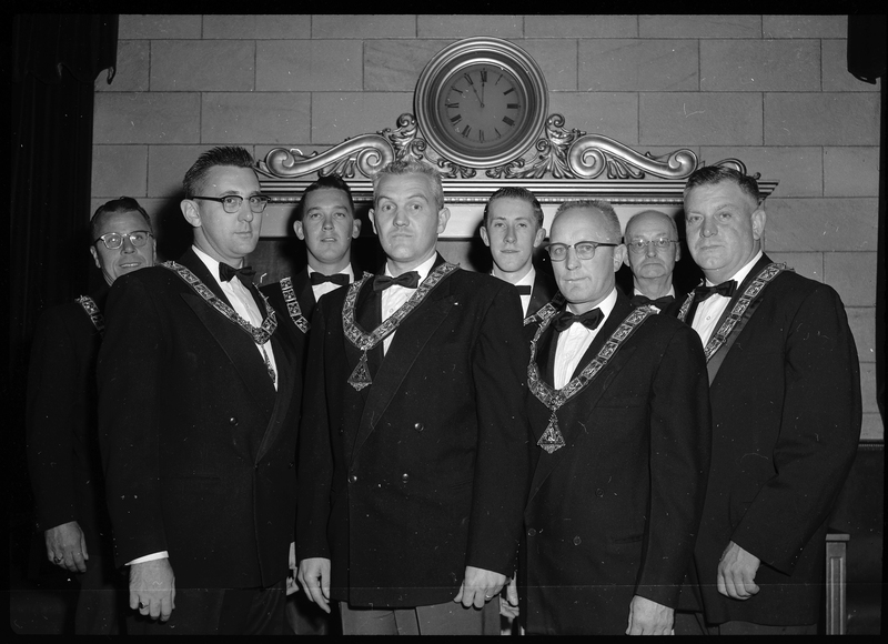 Photo of the eight men that are officers for the Benevolent and Protective Order of Elks. They are all wearing nice suit jackets and matching pendants while making serious faces. Most of the men are looking at the camera with a couple looking other places.