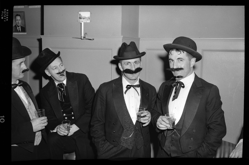 Photo of four men wearing fake mustaches, suits, and hats pose at a Rotary Club Party. They are also holding glasses.