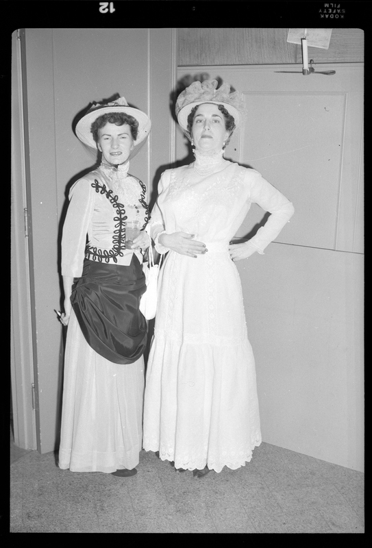 Two women in formal dresses and hats pose together for a picture while attending a Rotary Club party.
