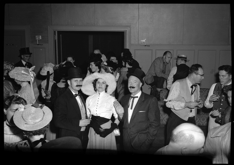 A group of people attending the Rotary Club party, with three people as the main subject of the photo. The two men in the subject are both dressed nicely while wearing hats and presumably fake mustaches, and the woman between them is wearing a fancy dress and hat. Almost everyone pictured in the background are dressed nicely and many are holding drinks.