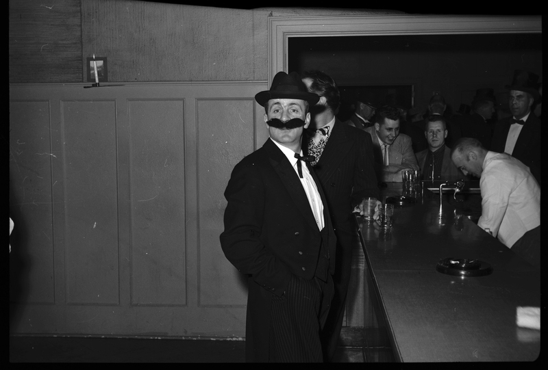 Photo of a man at a Rotary Club party. He is well dressed and wearing a fake mustache while standing at a bar. There are a few people visible in the background.