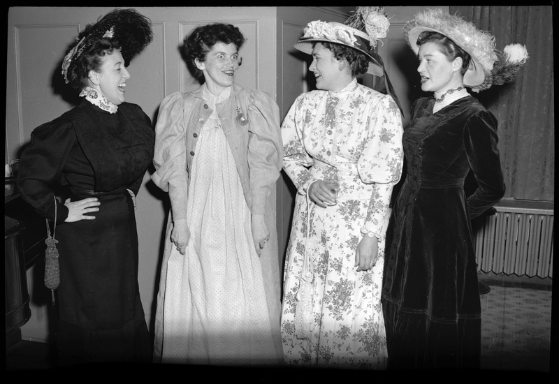Four women standing together at a Rotary Club party. One of the women is making a face and the other three are laughing at her. They are all wearing fancy dresses.