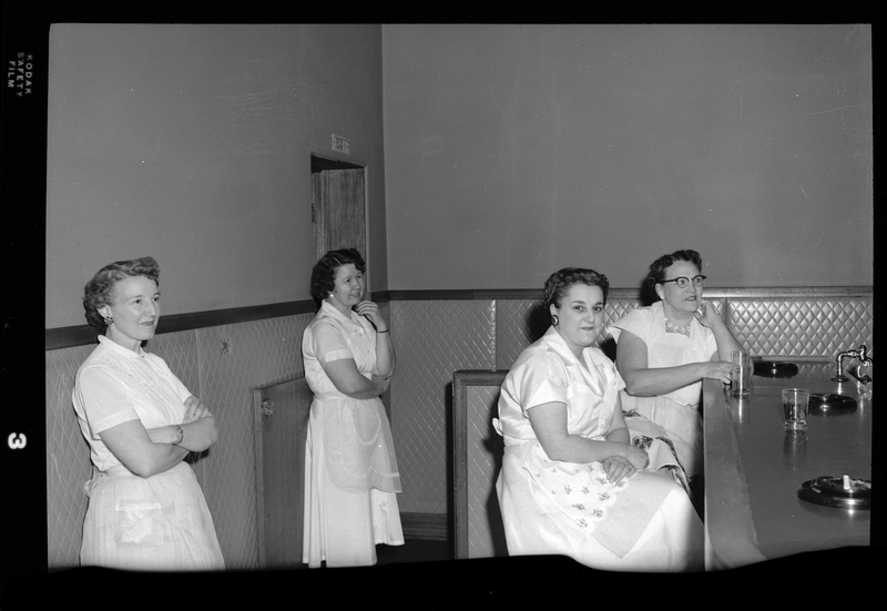 Four women wearing aprons at the Rotary Club party. Two women are sitting at the bar and the other two are standing behind them. One woman is looking at the camera.
