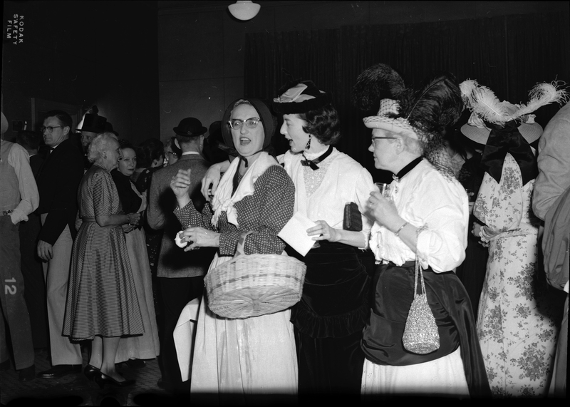 Photo of several women at a Rotary Club Party. Three women are centered in the photo with one of them looking at the camera. The women and the people in the background are formally dressed.