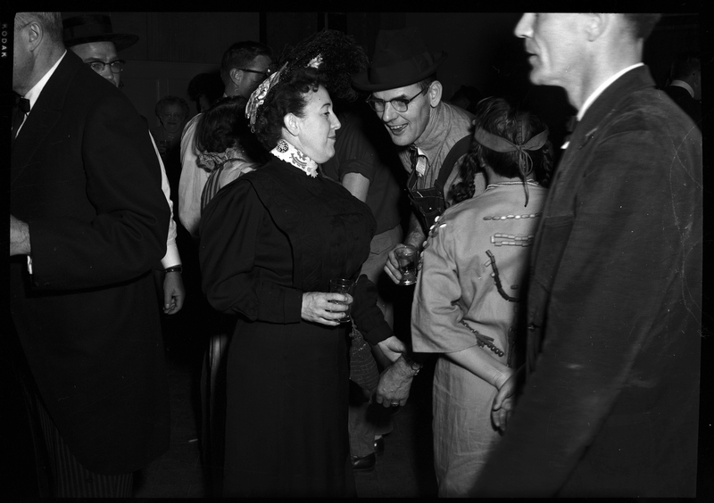 Candid photo of a woman reaching out for a man in a crowd of people at a Rotary Club party. She is wearing a dress and holding a drink. There are several people surrounding both the man and woman.