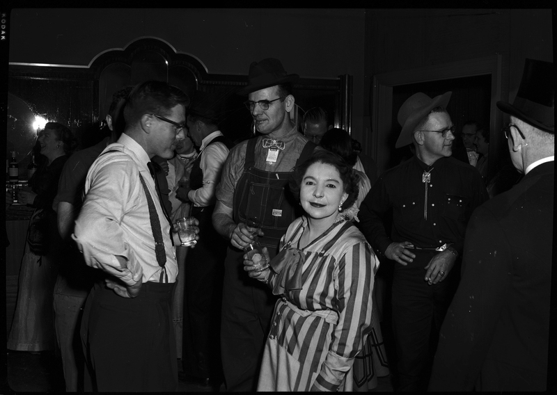 Several men stand together while talking with a woman standing in front of them, looking directly at the camera. They are all dressed nicely and holding drinks. 