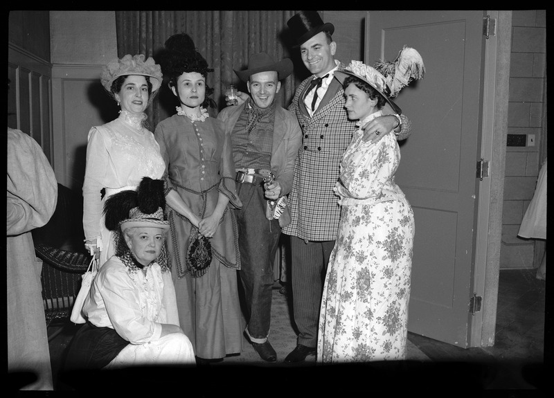 Group of men and mostly women posing for a photo together at a Rotary Club party. The women are all in fancy dresses and the men are wearing costumes. Most are looking at the camera and one man is pointing a (presumably) fake gun at the camera.