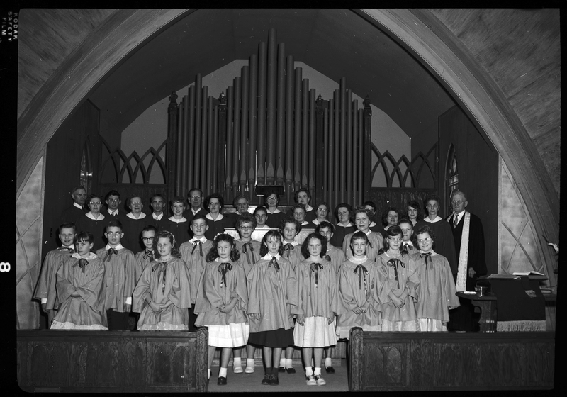 Staged photo of a church choir, possibly Methodist. Men, women, and children are lined up in rows and wearing matching robes. They are standing in front of what is likely an organ.