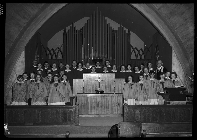 Staged photo of a church choir, possibly Methodist. Men, women, and children are lined up in rows with an open space in the middle showing what is most likely a Communion table. They are standing in front of what is likely an organ and wearing matching robes.