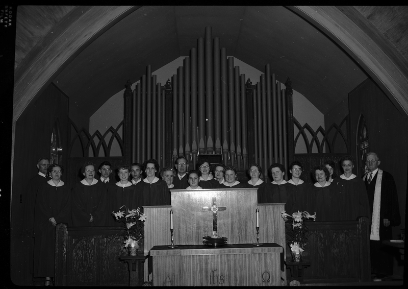 Staged photo of a church choir, possibly Methodist. Adult men and women are lined up behind what is likely a Communion table and in front of what is likely an organ. They are all wearing matching robes.