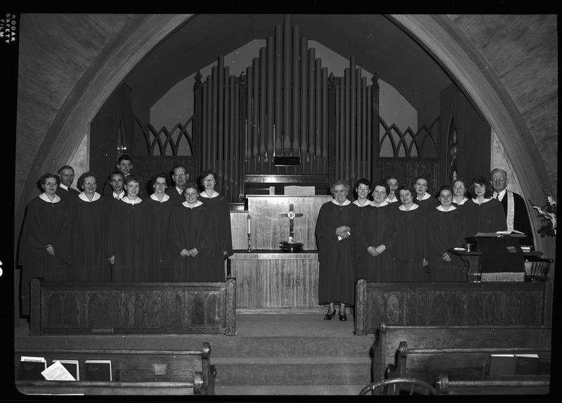 Staged photo of a church choir, possibly Methodist. Adult men and women are lined up behind what is likely a Communion table and in front of what is likely an organ. They are all wearing matching robes.