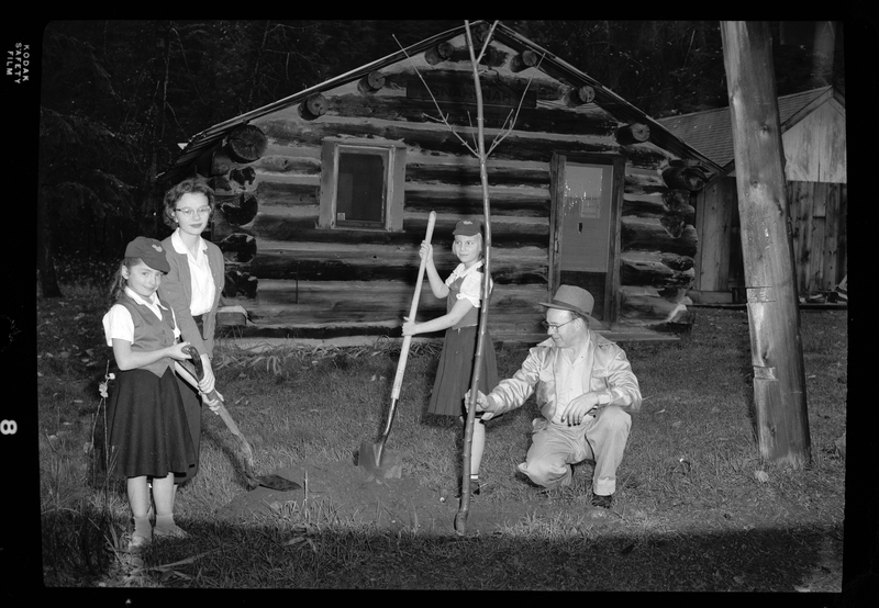 Photo of two young girls planting a tree with the help of a man and a woman. The two girls are wearing matching uniforms for Camp Fire Girls and they are both holding shovels. There is a log cabin behind them and they appear to be in a forest.