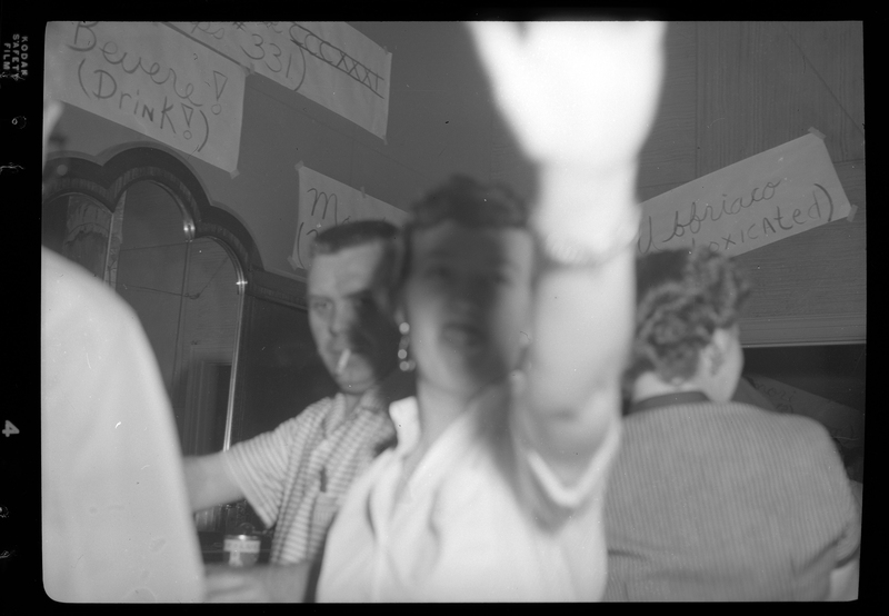 Photo of a woman holding her hand out to block the camera's view at a Rotary Club party. There is a man behind her with a cigarette in his mouth, a woman facing the other direction, and hand made signs on the wall are also visible.