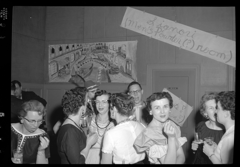 A candid photo of several women at a Rotary Club party. Most of them are talking to each other, but a few women are looking at the camera. Everyone is dressed nicely, and there are hand made signs on the walls behind them.