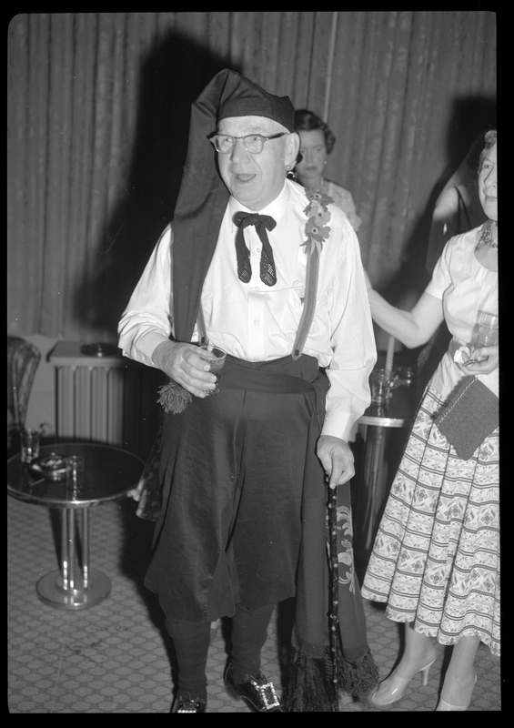 Photo of a man in a costume at a Rotary Club party. He is wearing a long hat and has flowers on his suspenders. He is holding a drink and there are women behind and next to him.