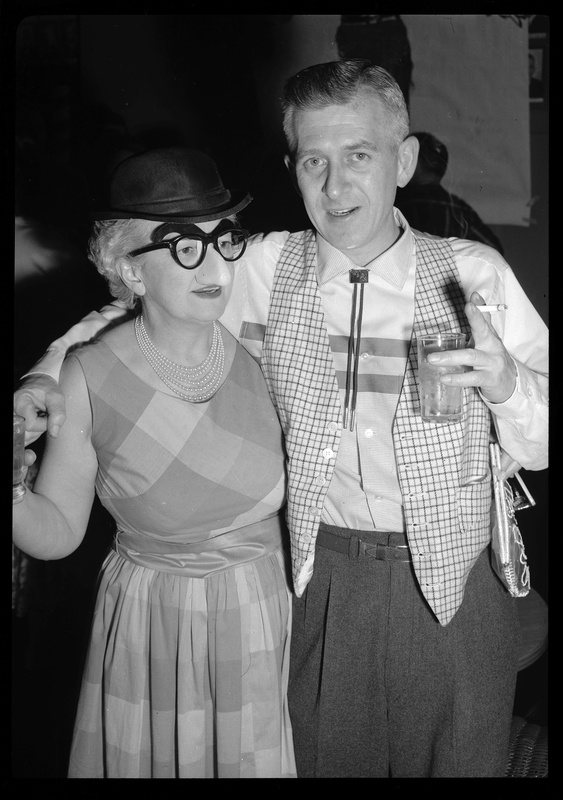 A man and a woman are standing together, the man looking at the camera while holding a drink and a cigarette in one hand. He has his arm wrapped around a woman's shoulders, who is wearing a fake glasses, eyebrows, and nose combination. They are at a Rotary Club party.