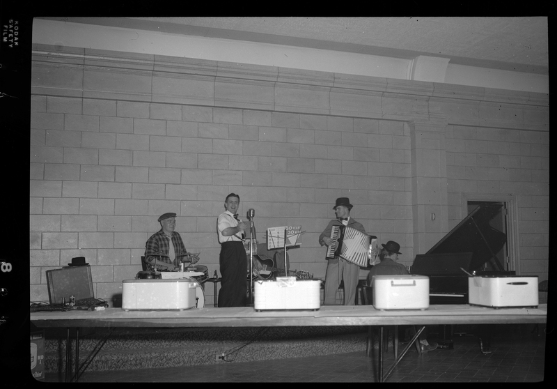 Photo of five men with instruments at the Rotary Club party. One man has a guitar, another has an accordian, a third has what appears to be a drum set, the fourth is seated at a piano, and the fifth man also has an accordion as well as the microphone in front of him.