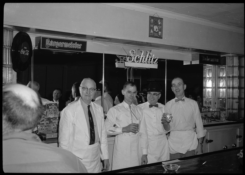 Four men stand behind a bar, wearing similar outfits, and posing for a photo. Two of the men are holding drinks. There is a neon sign on the wall behind them for Schlitz beer.
