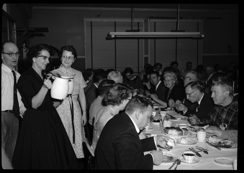 A woman is holding two jugs while standing behind a group of people dining at long tables. She appears to be trying to fill cups. There is a man and another woman standing near her.
