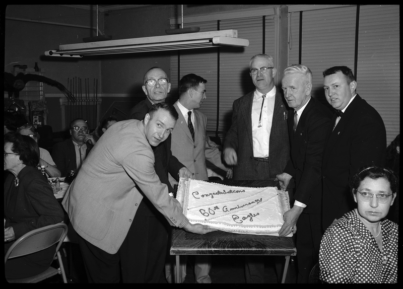 Six men stand around a decorated cake for the 60th anniversary of Fraternal Order of Eagles. A few of the men are trying to tilt the cake upwards to pose with it for the camera. People can be seen in the background eating.