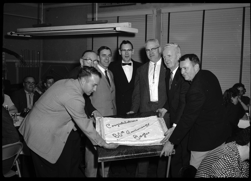 Seven men stand around a decorated cake for the 60th anniversary of Fraternal Order of Eagles. A few of the men are trying to tilt the cake upwards to pose with it for the camera. People can be seen in the background eating.