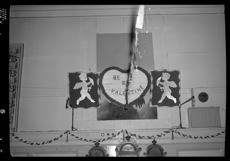 Photo of a hand made sign that reads "Be my Valentine" in the shape of a heart. On either side of the heart is the silhouette of cupid. These are hung together on a wall.