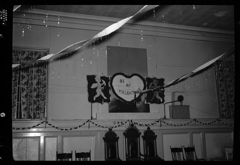 Photo of a hand made sign that reads "Be my Valentine" in the shape of a heart. On either side of the heart is the silhouette of cupid. These are hung together on a wall, and there are banners hung across the room.