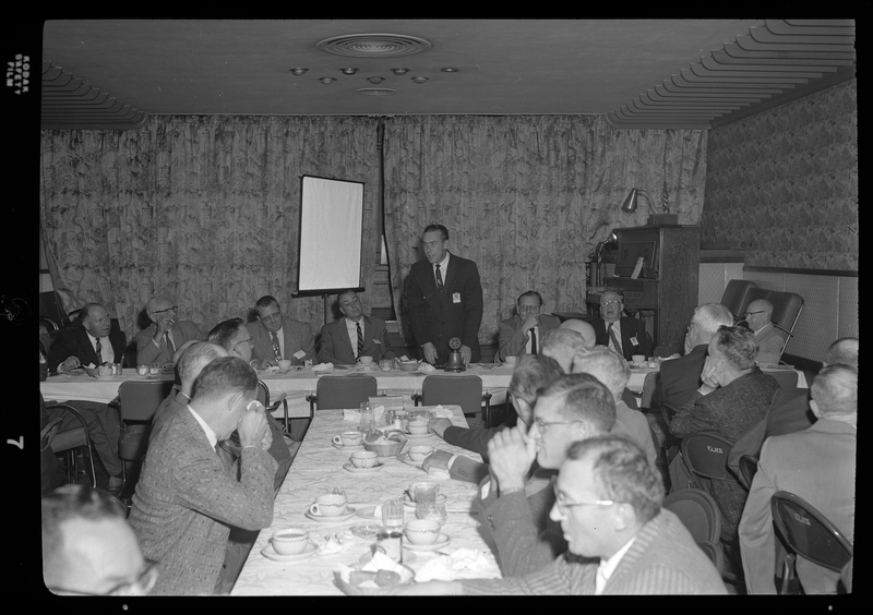 A group of people, seemingly all men, gather in a room for the Rotary Club in Spokane, Washington. Everyone is seated around tables with food on it, and they are all looking at a man standing in the back of the room.