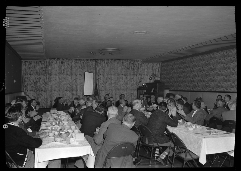 A group of people, seemingly all men, gather in a room for the Rotary Club in Spokane, Washington. Everyone is seated around tables with food on it.