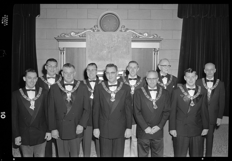 A group photo of the nine officers for the Benevolent and Protective Order of Elks. The men are standing in two rows wearing matching suit jackets, bow ties, and pendants. There is a painting of an elk behind them.