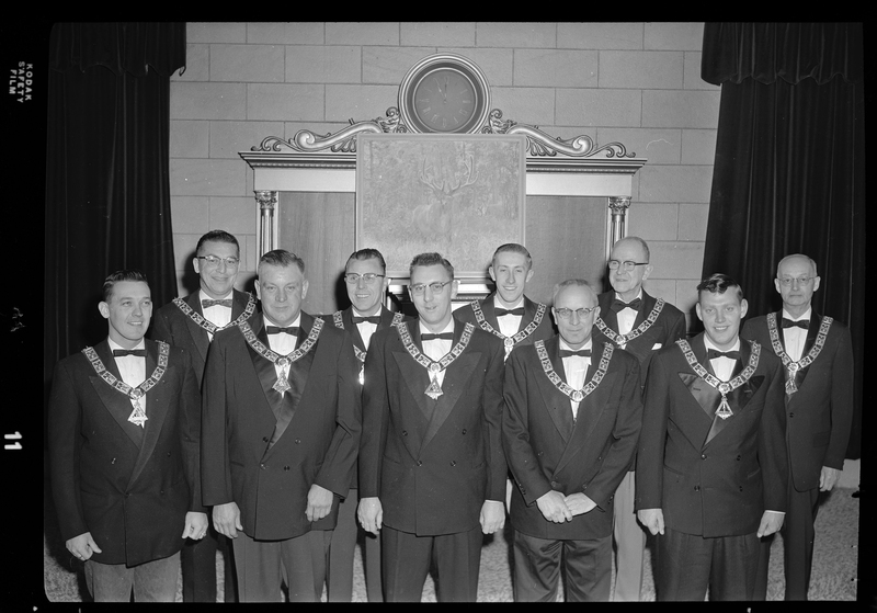 A group photo of the ten officers for the Benevolent and Protective Order of Elks. The men are standing in two rows wearing matching suit jackets, bow ties, and pendants. There is a painting of an elk behind them.