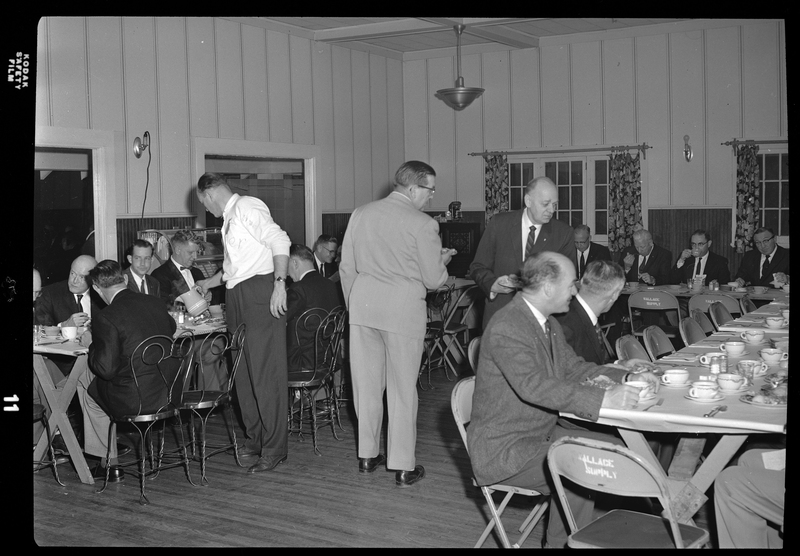 A group of well dressed men can be seen sitting at multiple tables in a room, with a few men standing in the middle. There are coffee cups set out on the tables and some of the men are drinking from them.