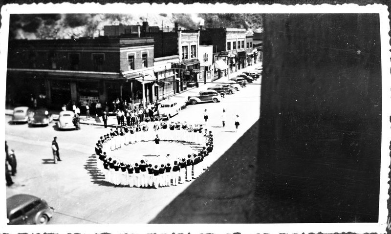 Damaged negative of the Wallace High School band performing on the corner of 6th and Cedar Street. The band can be seen standing in a circle around one person, with a small crowd watching. Buildings and cars are visible in the background.