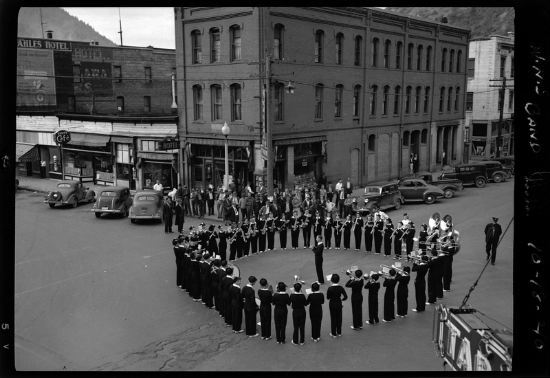 Photo of the Wallace High School band performing on the corner of 6th and Cedar Street in Wallace, Idaho. The band can be seen standing in a circle around the conductor in the middle of the street. There is a small crowd watching the band from the sidewalk, standing near buildings and cars.