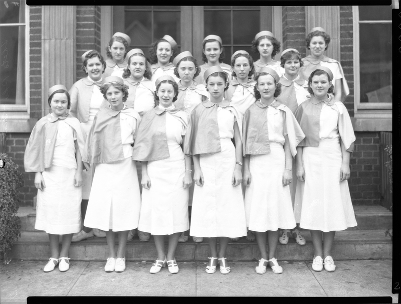Photo of sixteen women standing together in three rows. They are wearing matching outfits and standing on the steps of a building for the photo. Previously described as "Theta Rho Girl's Drill Team."