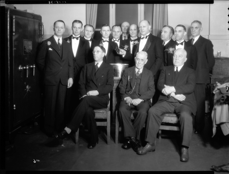 Fourteen men pose together for a photo at an event for the Benevolent and Protective Order of Elks, likely the Burning Mortgage and Banquet. Three of the men are seated in chairs with the rest of the men standing behind them.