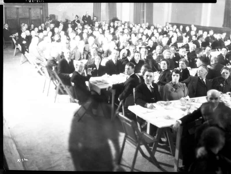 A large crowd of people seated at tables for an event by the Benevolent and Protective Order of Elks, likely the Burning Mortgage and Banquet. Some of the people's faces are washed out, but it looks as if everyone is looking at the camera for the photo. The shadow of a person can be seen walking in front of the camera.