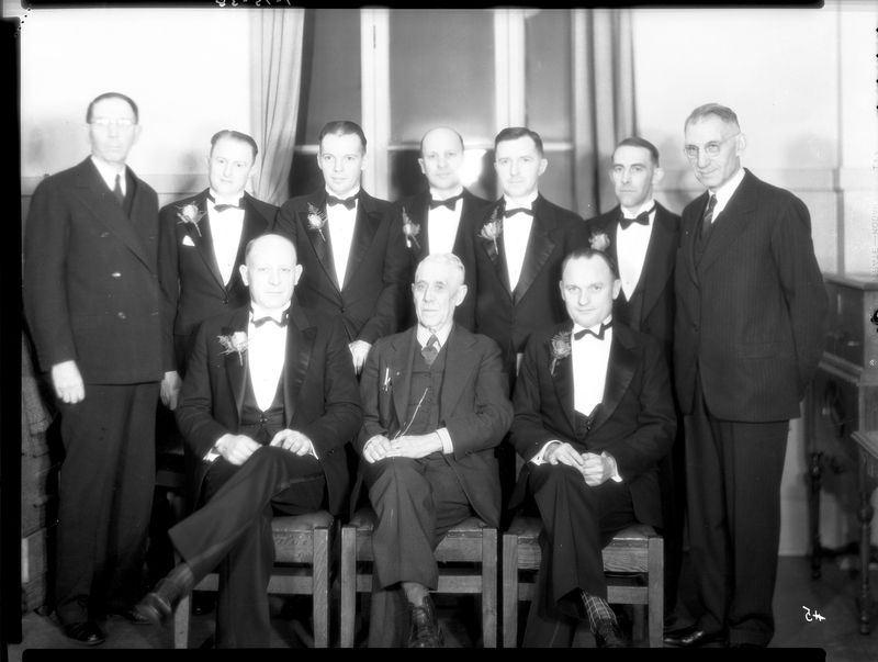 Photo of ten men posing for a photo together at a Benevolent and Protective Order of Elks event, likely the Burning Mortgage and Banquet. Three of the men are sitting while the others stand behind them. They are all dressed in suits and a few are wearing boutonnieres.