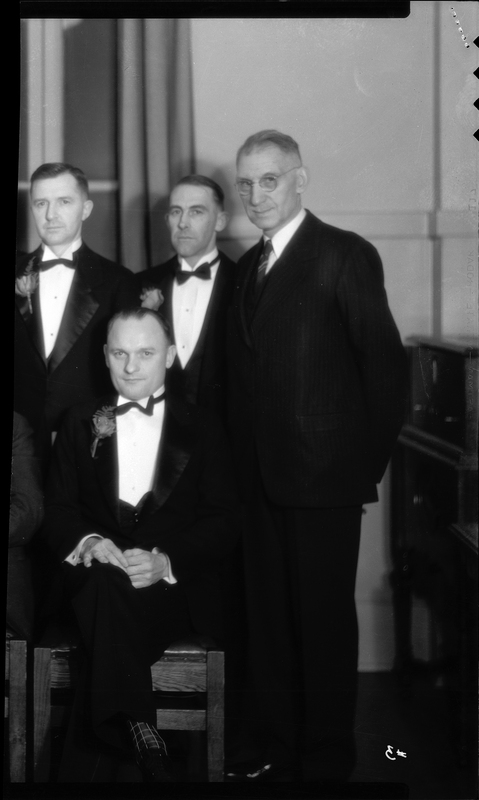 Photo of four men posing for a photo together at a Benevolent and Protective Order of Elks event, likely the Burning Mortgage and Banquet. One man is sitting while the others stand behind them. They are all dressed in suits and a few are wearing boutonnieres.
