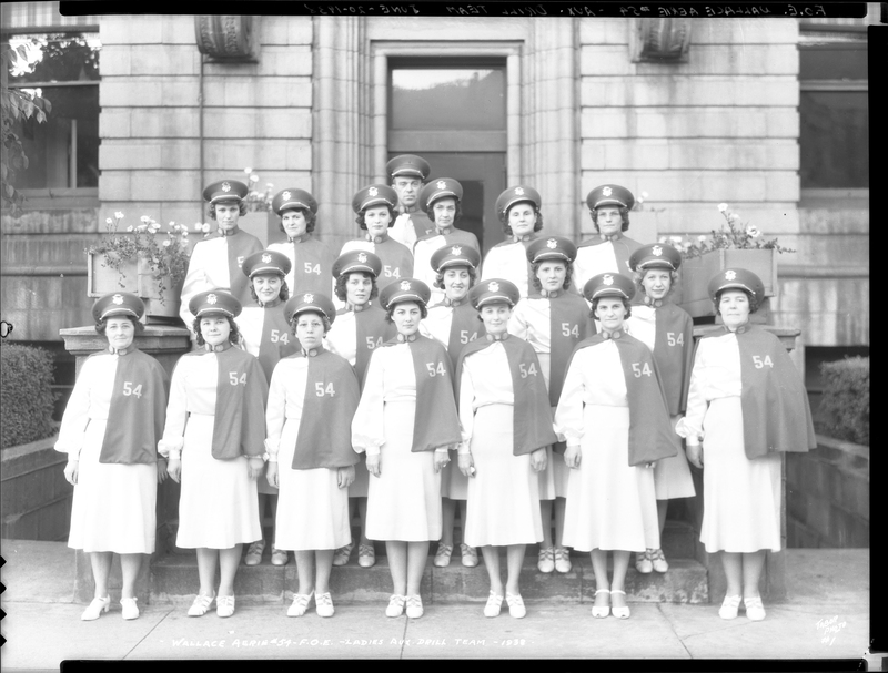 Group of women in matching uniforms stand in rows on the steps of a building for a photo. They are the Fraternal Order of Eagles ladies auxiliary drill team (Wallace Aerie #54).